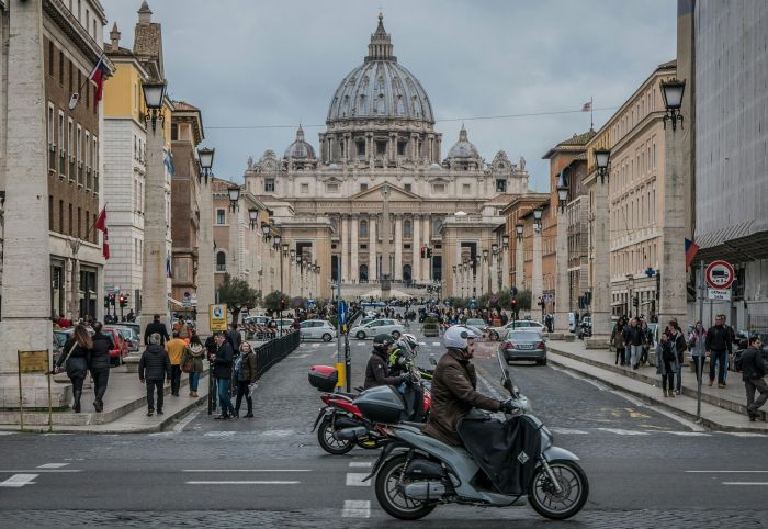Saint Peters Basilica's front facade and main square opens to a busy road with traffic and pedestrian.